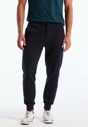 Mann trägt schwarze Jogginghosen mit Kordelzug, weiße Turnschuhe und ein dunkelgrünes T-Shirt vor weißem Hintergrund.