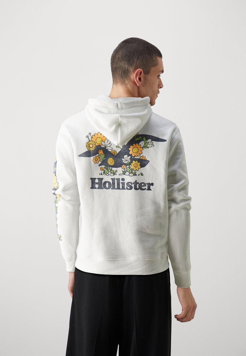 Mann in weißem Hoodie mit floralem Hollister-Logo auf dem Rücken und floralem Design am rechten Ärmel, der mit dem Rücken zu einer hellen, einfarbigen Hintergrund steht.