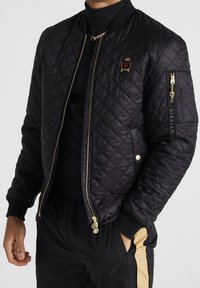Svart vadderad bomberjacka med en guldfärgade dragkedja, branding på ärmen och ribbade manschetter. Matchas med svarta byxor med gulddetaljer.