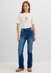 Camiseta de manga corta de un color beige claro con diseño gráfico en verde, combinada con unos vaqueros acampanados de color azul oscuro. Unos zapatos marrones completan el conjunto.