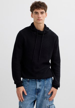 Jersey de punto negro con cuello vuelto y cordones. Presenta mangas largas y tejido texturizado. Combinado con vaqueros cargo de color azul claro.