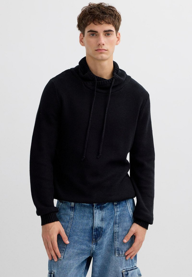 Jersey de punto negro con cuello vuelto y cordones. Presenta mangas largas y tejido texturizado. Combinado con vaqueros cargo de color azul claro.