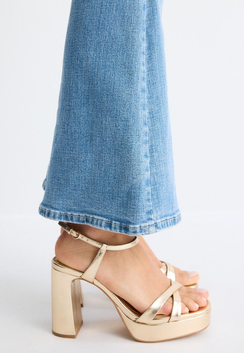 Femme portant un jean denim bleu clair et des sandales dorées à talons hauts avec des lanières à la cheville, debout sur une surface blanche.