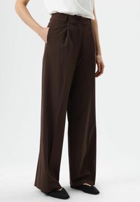 Pantalon large marron en tissu lisse, doté d'une taille haute, de plis et de poches latérales, associé à des chaussures noires sans lacets.