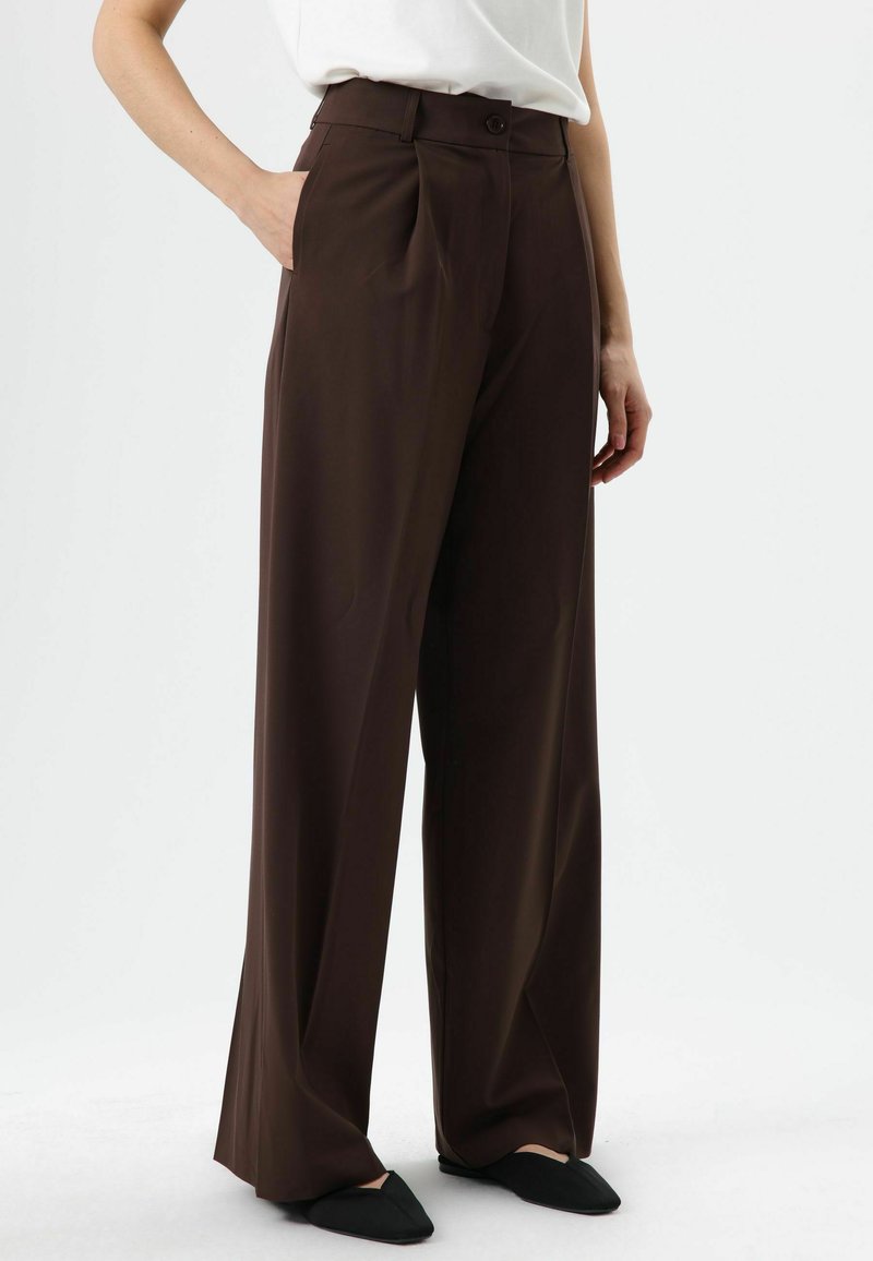 Pantalon large marron en tissu lisse, doté d'une taille haute, de plis et de poches latérales, associé à des chaussures noires sans lacets.