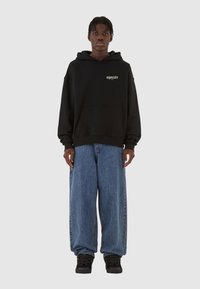 Svart hettegenser med frontlomme og logo, kombinert med baggy blå denimjeans. Svart sneakers fullfører antrekket.