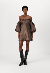 Barna selyem off-shoulder ruha, dús ujjakkal és ráncolt melle résszel, térd felett magassarkú fekete csizmával viselve, minimalista háttérrel.