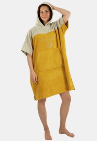 Kapuzenhandtuch-Poncho in Senfgelb und Beige, aus Frottiermaterial, mit einer Fronttasche und gesticktem Logo. Weiches, strukturiertes Material.