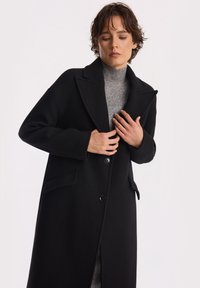 Osoba z krótkimi włosami, ubrana w długi czarny płaszcz na szary sweter z golfem, poprawiająca płaszcz obiema rękami.