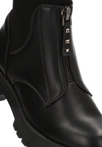 Botín negro de cuero hasta el tobillo con detalles acanalados, cremallera frontal y acentos de tachuelas metálicas; suela robusta para estabilidad y agarre.