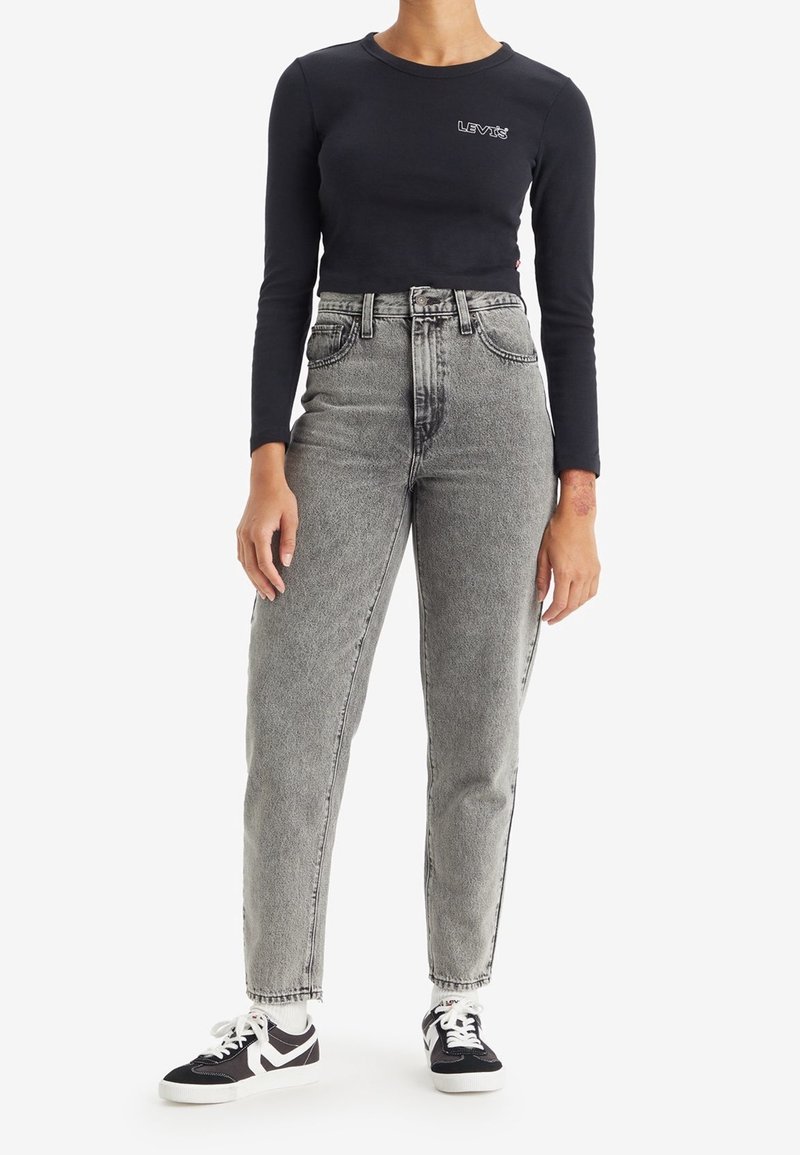 Personne portant un haut Levi's noir à manches longues, un jean gris taille haute, des chaussettes blanches et des baskets noires et blanches, debout devant un fond blanc.