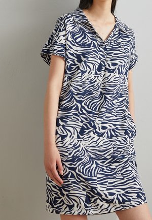 Frau trägt ein kurzärmeliges Kleid mit Kragen und einem abstrakten Zebra-Streifenmuster in Blau und Weiß, steht vor einem grauen Hintergrund.