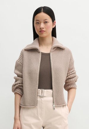 Vrouw draagt een beige gebreid vest met rits en opgerolde mouwen, een bruin geribd topje en lichtbeige hoge taille broek met riem.