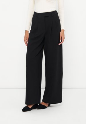 Femme portant un pantalon noir taille haute à jambes larges avec des plis, associé à des ballerines noires pointues et un haut blanc à manches longues.