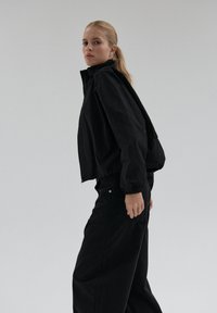 Młoda kobieta z blond kucykiem, ubrana w czarną kurtkę i szerokie czarne spodnie, stojąca na jednolitym jasnoszarym tle.