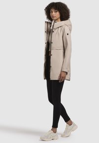 Beige waterdichte jas met een capuchon, drukknoopsluiting en een zwarte voering. Gedragen over een zwarte top en gecombineerd met zwarte leggings en witte sneakers.