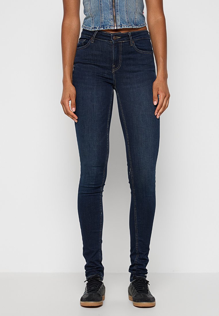 Kvinna i mörkblå skinny jeans, svarta sneakers med gummisulor, och en ärmlös kort denimjacka, som står mot en vit bakgrund.