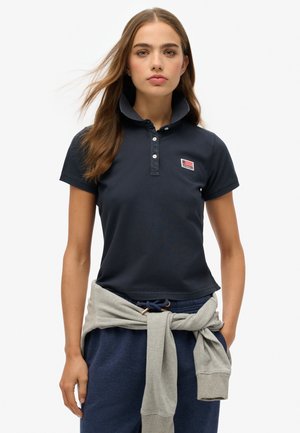 Joven mujer que lleva una camisa polo azul marino con un parche de la bandera americana y una sudadera gris atada alrededor de su cintura, de pie contra un fondo blanco.