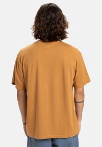 Man met krullend bruin haar, gekleed in een eenvoudige mosterdkleurige t-shirt en blauwe broek, gezien van de achterkant tegen een witte achtergrond.