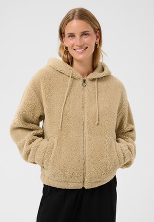 Young woman smiling with hands in pockets wearing beige zip-up fleece hoodie and black pants against plain background.
