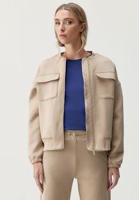 Beige bomberjacka med en slät finish, som har två framfickor och dragkedjestängning. Matchad med en blå ribbad topp och matchande beige byxor.