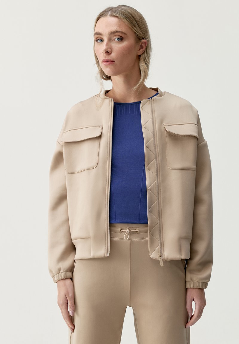 Beige bomberjacka med en slät finish, som har två framfickor och dragkedjestängning. Matchad med en blå ribbad topp och matchande beige byxor.