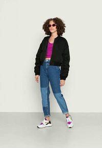 Casaco bomber preto, top crop roxo, calças de denim bicolores, sapatilhas brancas com detalhes roxos e verdes, óculos de sol redondos, cabelo crespo.