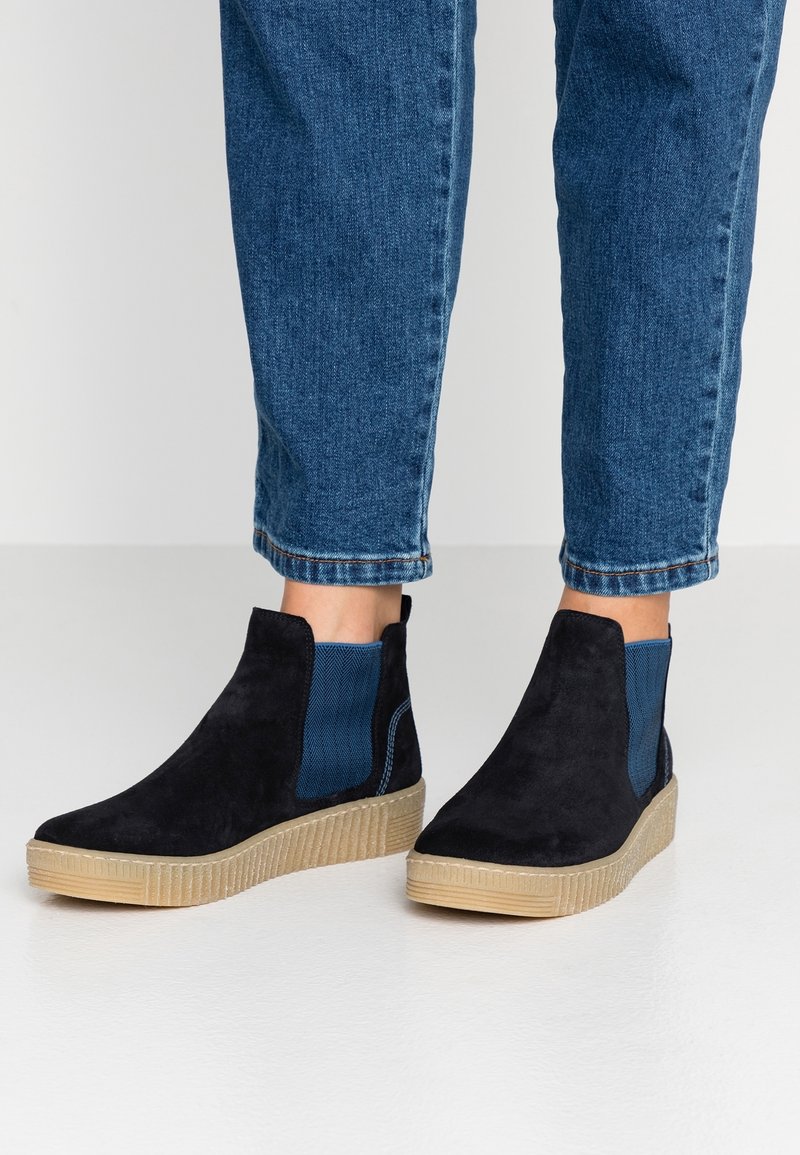 Personne portant des bottines en daim noir avec des semelles en caoutchouc beige et des panneaux latéraux élastiques bleus, associées à un pantalon en denim bleu.