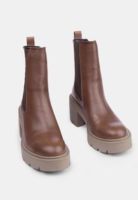 Botas de cuero marrón hasta el tobillo con paneles laterales elásticos, tacones de plataforma beige gruesos y puntas redondeadas. Textura suave y diseño minimalista.