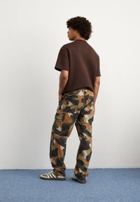 Ruskea lyhythihainen paita, maastokuvioiset cargo-housut, joissa on vihreitä, ruskeita ja mustia laikkuja, sekä raidalliset vihreät ja valkoiset matalaotsaiset lenkkarit.