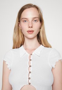 Camisola branca de malha com gola, mangas curtas e detalhe de botões. Apresenta uma bainha assimétrica e um design com recorte frontal com botões redondos.