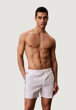 Homme torse nu aux cheveux courts noirs portant un short de bain blanc avec des bandes logo sur les côtés, debout devant un fond clair uni.