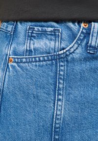 Prim-plan cu buzunarul unei perechi de blugi din denim albastru, cu cusături vizibile, nituri din cupru și o cămașă neagră băgată în talie.