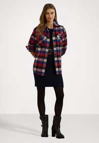 Röd, mörkblå och kremrutigt flanellskjorta med två bröstfickor, bärs över en mörkblå stickad klänning och matchas med svarta ankelstövlar.