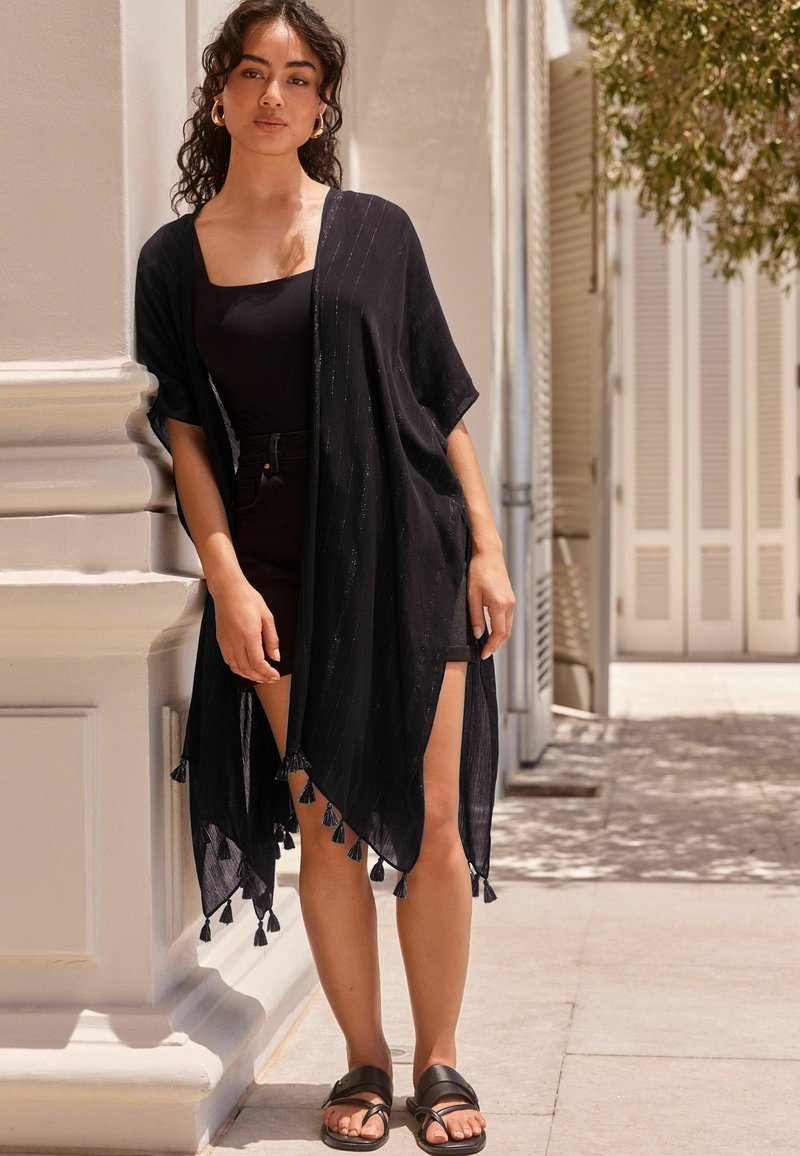 Czarny kimono w stylu narzutki wykonany z lekkiego materiału, z teksturowanymi pasami i frędzlami na dolnej krawędzi, noszony na czarnym stroju.