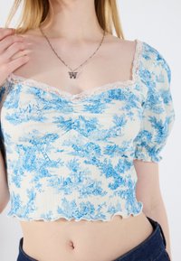 Lyseblå blomsterprint crop top med puffærmer, blonder langs udkanten af halsen og en rynket kant. Materialet ser tekstureret ud med en let stræk.