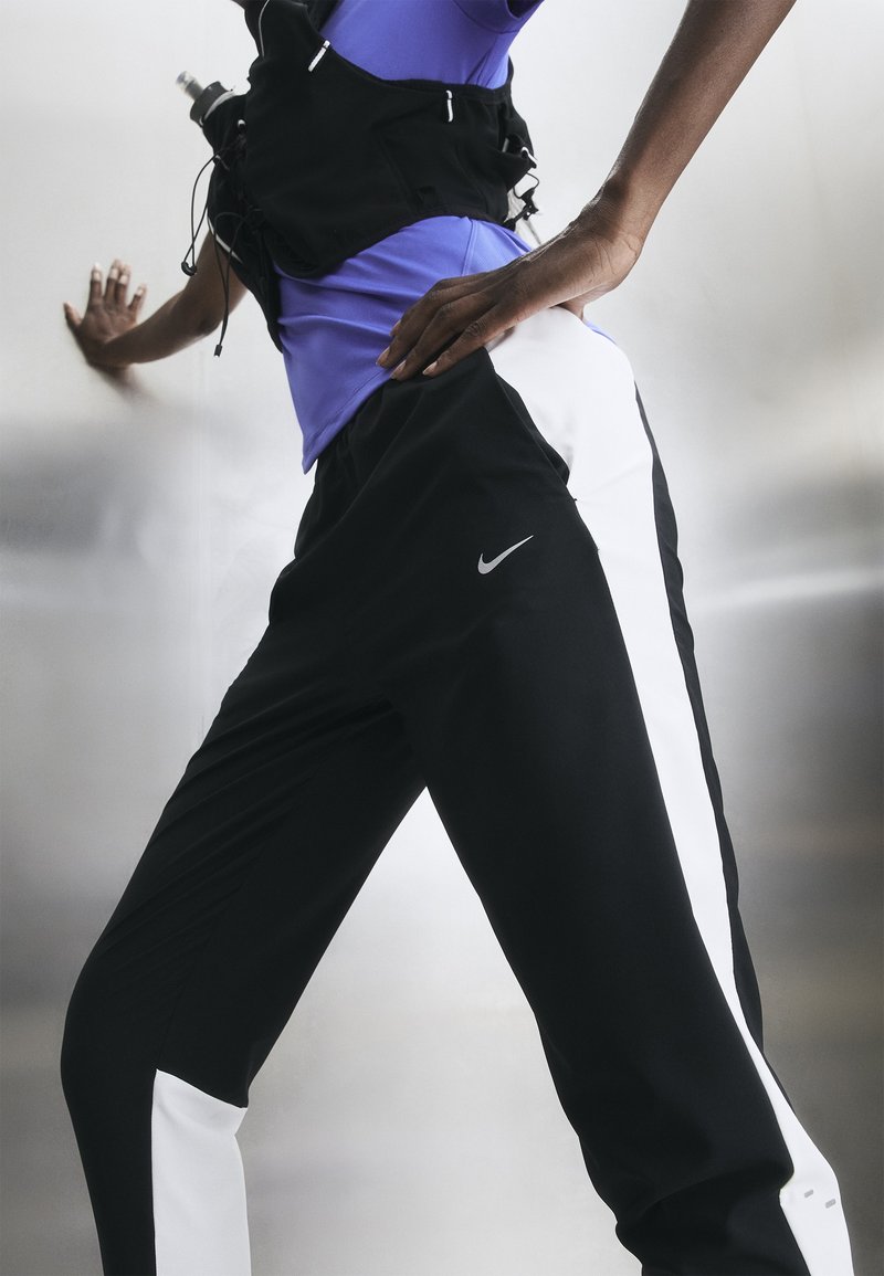 Άτομο με μαύρη και άσπρη φόρμα Nike και μωβ τοπ, ακουμπισμένο σε αντανακλαστική επιφάνεια με το ένα χέρι στη μέση.