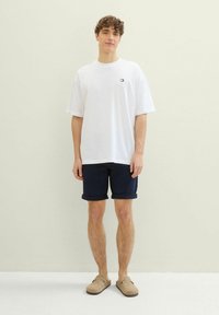 Biała bawełniana koszulka z krótkim rękawem i małym logo, w połączeniu z granatowymi szortami i beżowymi butami wsuwanymi o teksturowanej powierzchni.