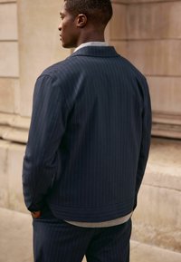 Mørkeblå stribet jakke med spredt krave, afslappet pasform og tekstureret grå turtleneck under. Glat stof med subtil glans.