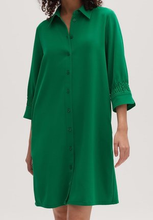 Woman wearing knee-length green button-up dress with long sleeves and shirred cuffs, standing against plain background.