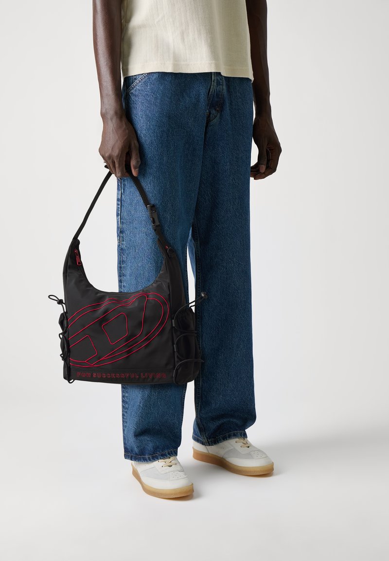 Sac à bandoulière noir avec des coutures rouges, présentant un design courbé, une bandoulière réglable et des poches latérales. Le texte indique "POUR UNE VIE RÉUSSIE."