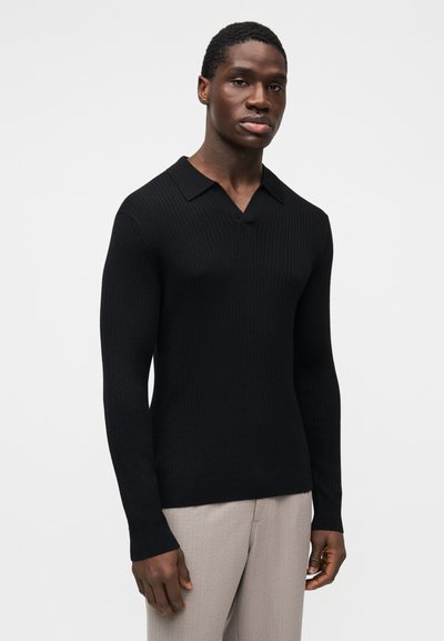 Pull noir côtelé avec col en V et col rabattu, manches longues ajustées et finition texturée. Porté avec un pantalon gris clair.