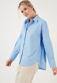 Light blue button-up shirt with a collar, long sleeves, and a slightly curved hem. Made from woven fabric with a smooth texture.