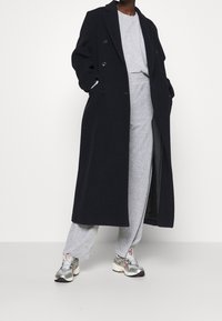 Abrigo largo negro de lana con botones de doble botonadura, usado sobre una sudadera gris y pantalones grises a juego. Par de zapatillas metálicas.