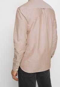 Beige langarmshirt met een zachte textuur, knoopmanchetten en een lijfje ontwerp. Beschikt over een licht gebogen zoom en een rugplooidetail.