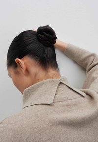 Frau mit dunklem Haar in einem gedrehten Dutt, die einen beigen Pullover mit hohem Kragen trägt und mit erhobenem Arm vor einer hellgrauen Wand steht.