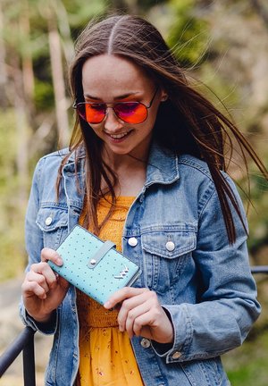 Türkisfarbene Geldbörse mit Punkten und Druckknopfverschluss. Verfügt über ein metallisches Akzentelement. Modell trägt eine Jeansjacke und ein oranges Oberteil.
