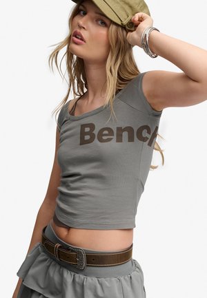 Joven mujer que lleva una camiseta gris corta con el texto "Bench", falda con volantes, cinturón marrón, pulseras y una gorra verde oliva, posando con la mano en la gorra.