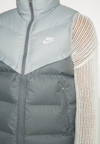 Grå pufferväst med hög krage, dragkedja fram och sidficka; har ett ljusgrått chevronmönster och kontrasterande vita nätärm.