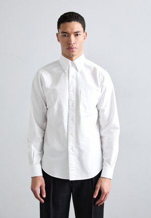 Weißes Baumwollhemd mit langen Ärmeln, Button-Down-Kragen, Brusttasche und geradem Saum. Kombiniert mit dunklen, taillierten Hosen.